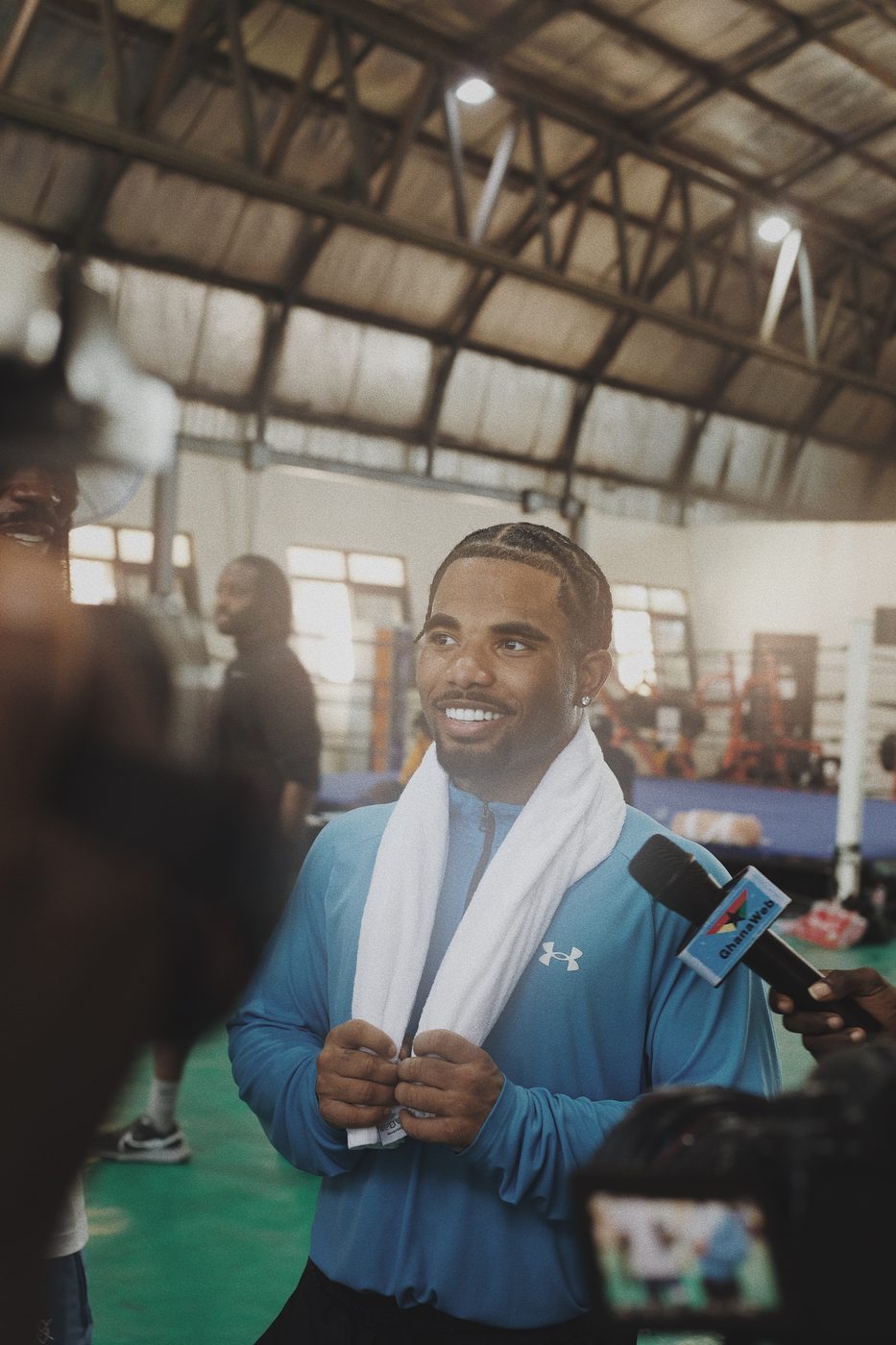 An athlete in a blue training top, a towel round his neck, smiling as he is interviewed at a boxing gym