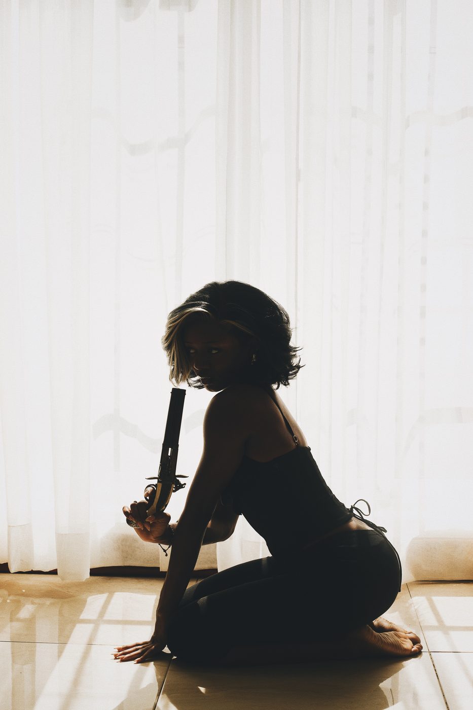 A woman kneeling in silhouette against a bright curtained window, holding an ornate long-barrelled pistol